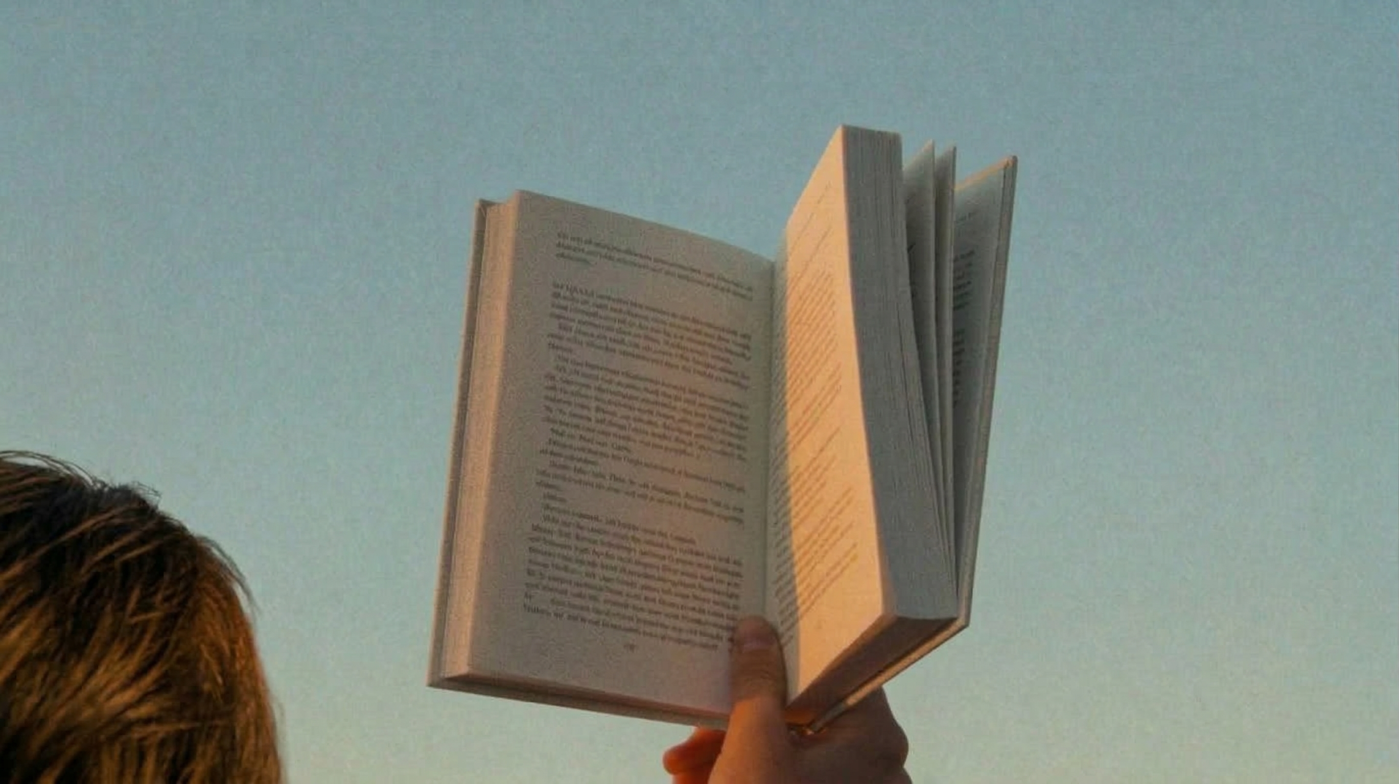 A hand holding an open book up against the sky at golden hour